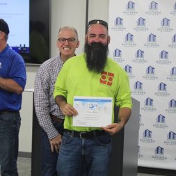 Man with a beard holding a certificate, smiling with three other men in a room with a projector screen and a backdrop with logos.