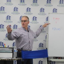 Man in glasses and checked shirt gesturing towards a whiteboard with "ford 8," "chev 14," and "$80,000" written on it, in a room with branded banners in the background.